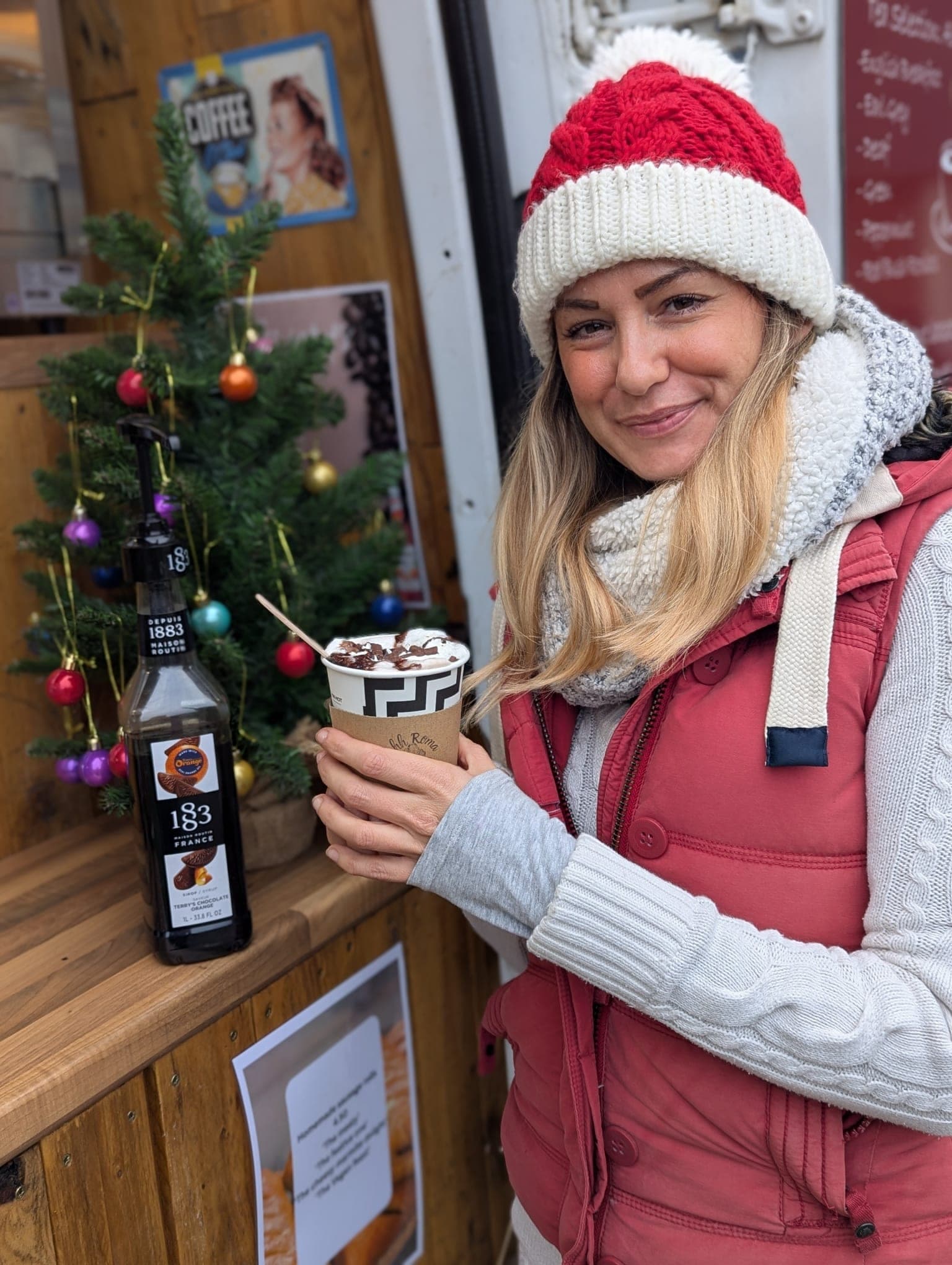 Happy customer holding an Ahh Roma hot chocolate with cream beside a 1883 syrup bottle and Christmas tree