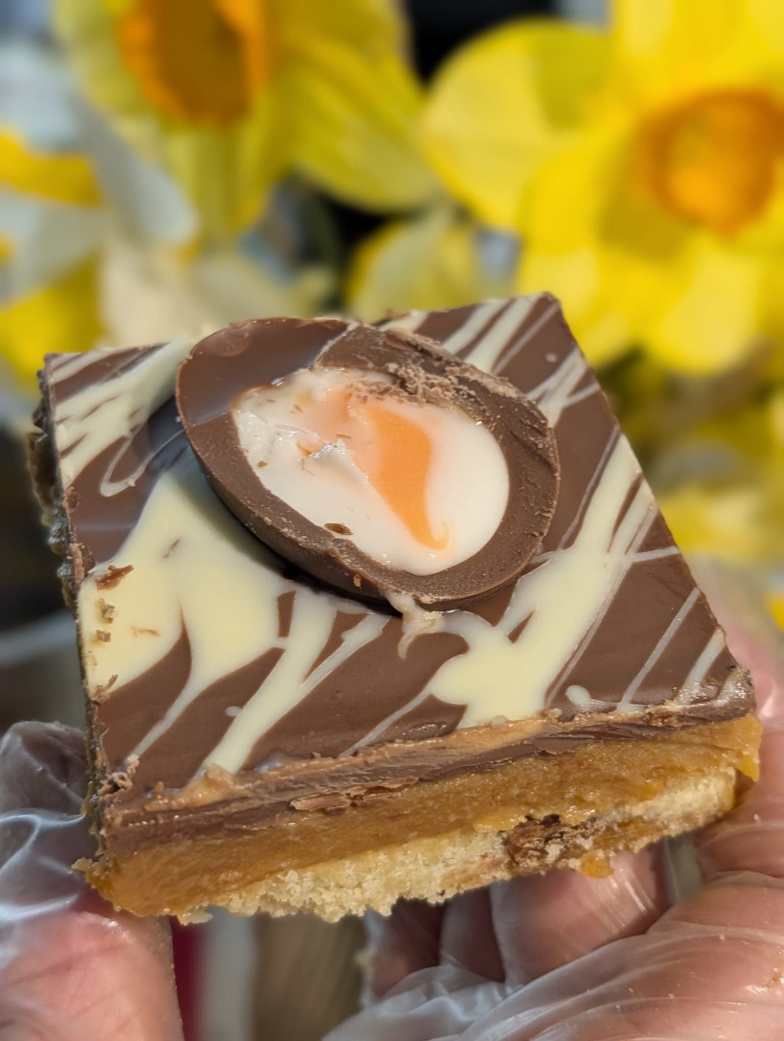 Millionaire's shortbread topped with a halved Creme Egg, held in a gloved hand with daffodils in the background